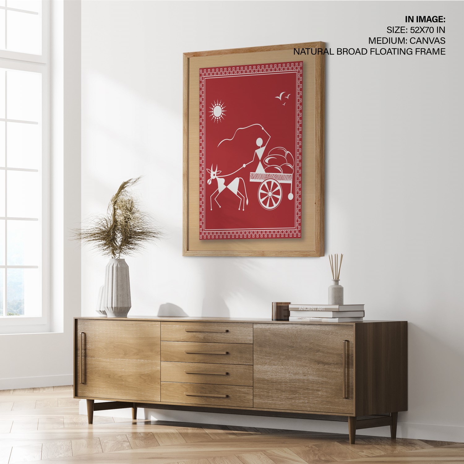 Large Warli art canvas in natural broad frame hung above a wooden credenza — white stick figures with bullock cart on terracotta red, lifestyle room mockup